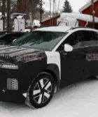 Vehículo camuflado en pruebas, estación de carga, nieve, edificios rojos.