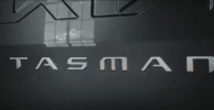 Logotipo metálico con la palabra "TASMAN" en un fondo oscuro.