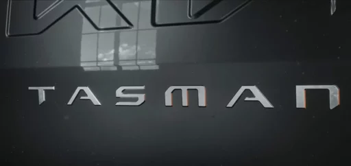 Logotipo metálico con la palabra "TASMAN" en un fondo oscuro.