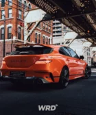 Coche naranja deportivo en un entorno urbano, bajo un puente elevado.