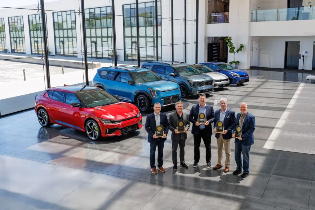 Cinco hombres sostienen trofeos delante de varios automóviles expuestos.