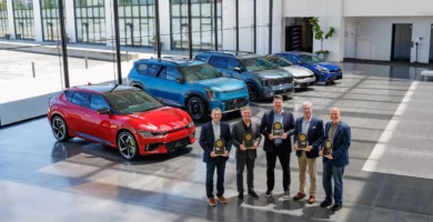 Cinco hombres sostienen trofeos delante de varios automóviles expuestos.