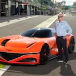 Hombre posando junto a un auto deportivo naranja en un circuito.